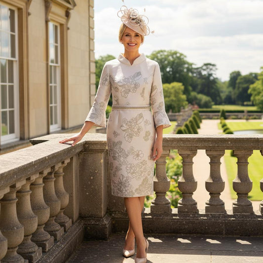 Woman in a formal Veni infantino gold mother of the bride dress and fascinator standing on a balcony with a scenic background. Signature Dress.