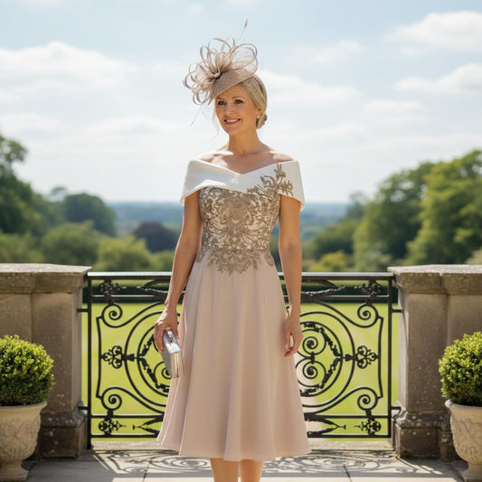 Woman in an elegant veni infantino taupe mother of the bride bardot dress with a decorative hat standing on a balcony with a scenic background. Signature Dress.