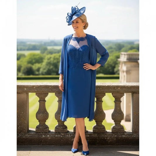Woman in a Veni Infantino blue mother of the bride dress and jacket and matching blue fascinator hat standing on a stone balcony with a garden background. Signature Dress.
