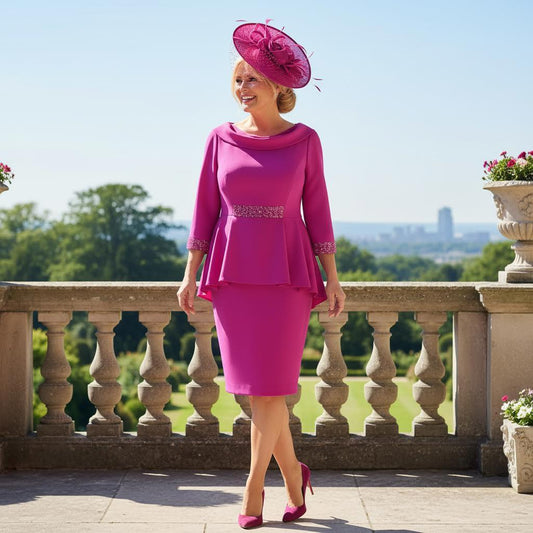 Woman in a plus size Veromia pink mother of the bride dress and matching pink fascinator hat standing on a balcony with a scenic background. Signature Dress.