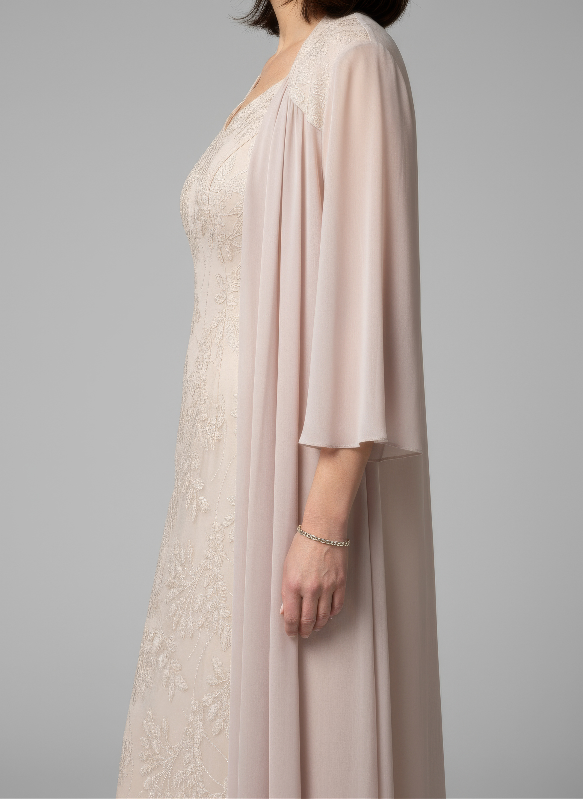 Woman wearing a long, light champagne dress with a matching cardigan against a gray background