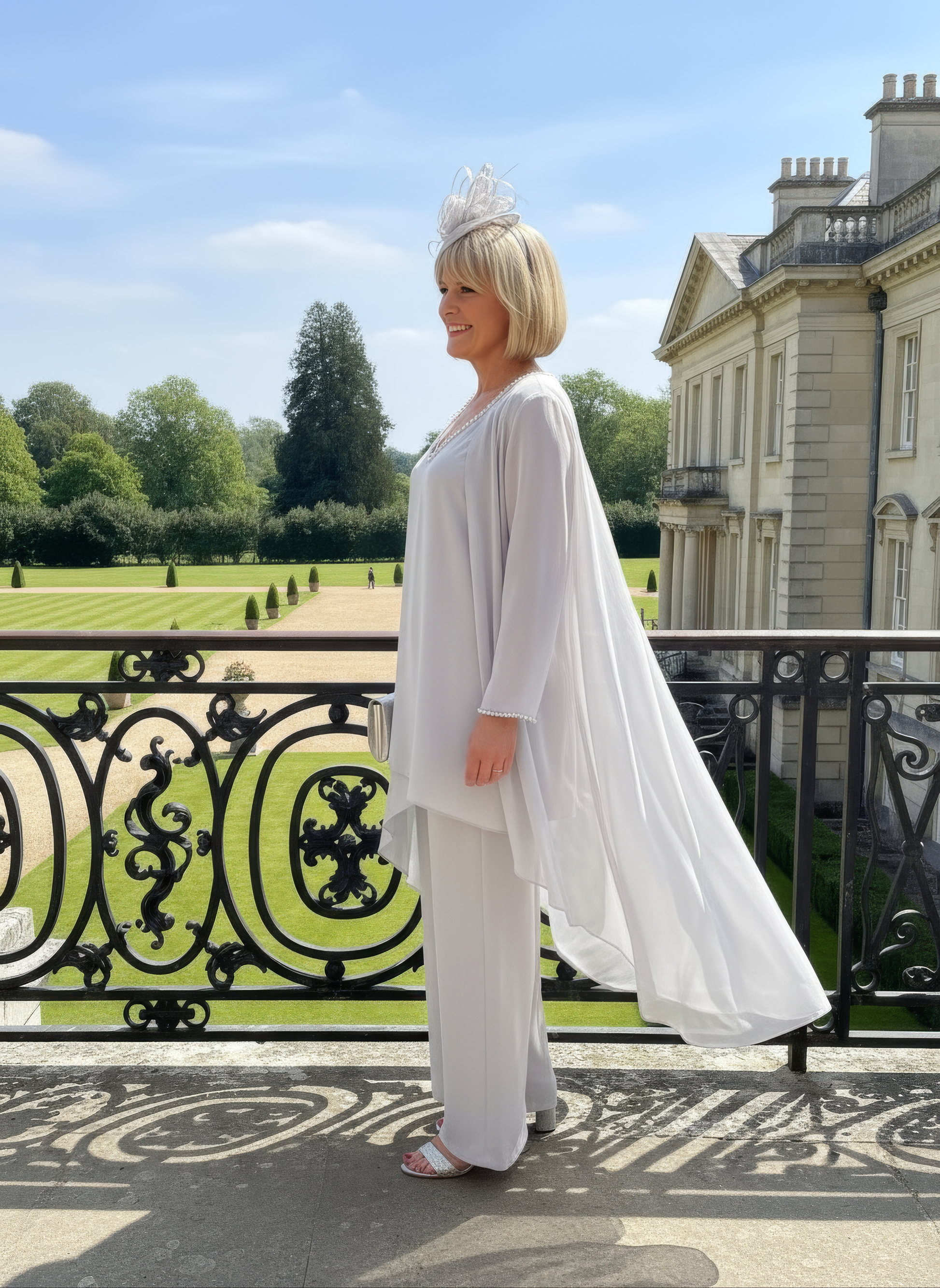 Woman in a silver mother of the bride trouser suit outfit make by veromia, standing in front of a large building with a garden.