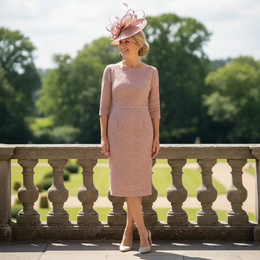 Woman in a veromia blush pink mother of the bride dress and pink fascinator hat standing on a stone balcony ready for a wedding. Signature Dress.