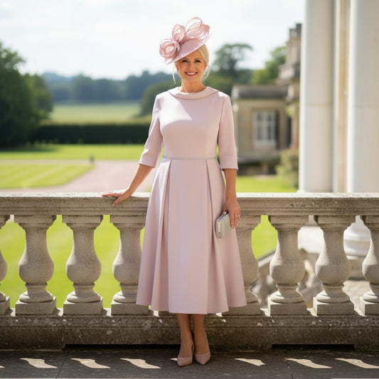 Woman in a Veromia light pink mother of the bride dress and hat standing on a balcony, the dress is midi length with 3/4 sleeves.