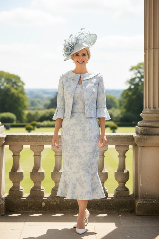 Woman in a light blue dress and jacket with a matching hat standing on a balcony with a scenic background.
