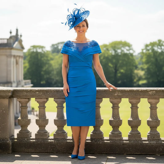 Woman in a blue mother of the bride dress and blue fascinator hat standing on a stone balcony with greenery in the background