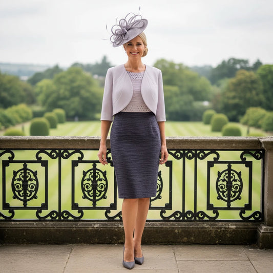 Woman in formal grey mother of the bride dress and jacket with mauve fasciantor hat standing in front of decorative railing with garden view
