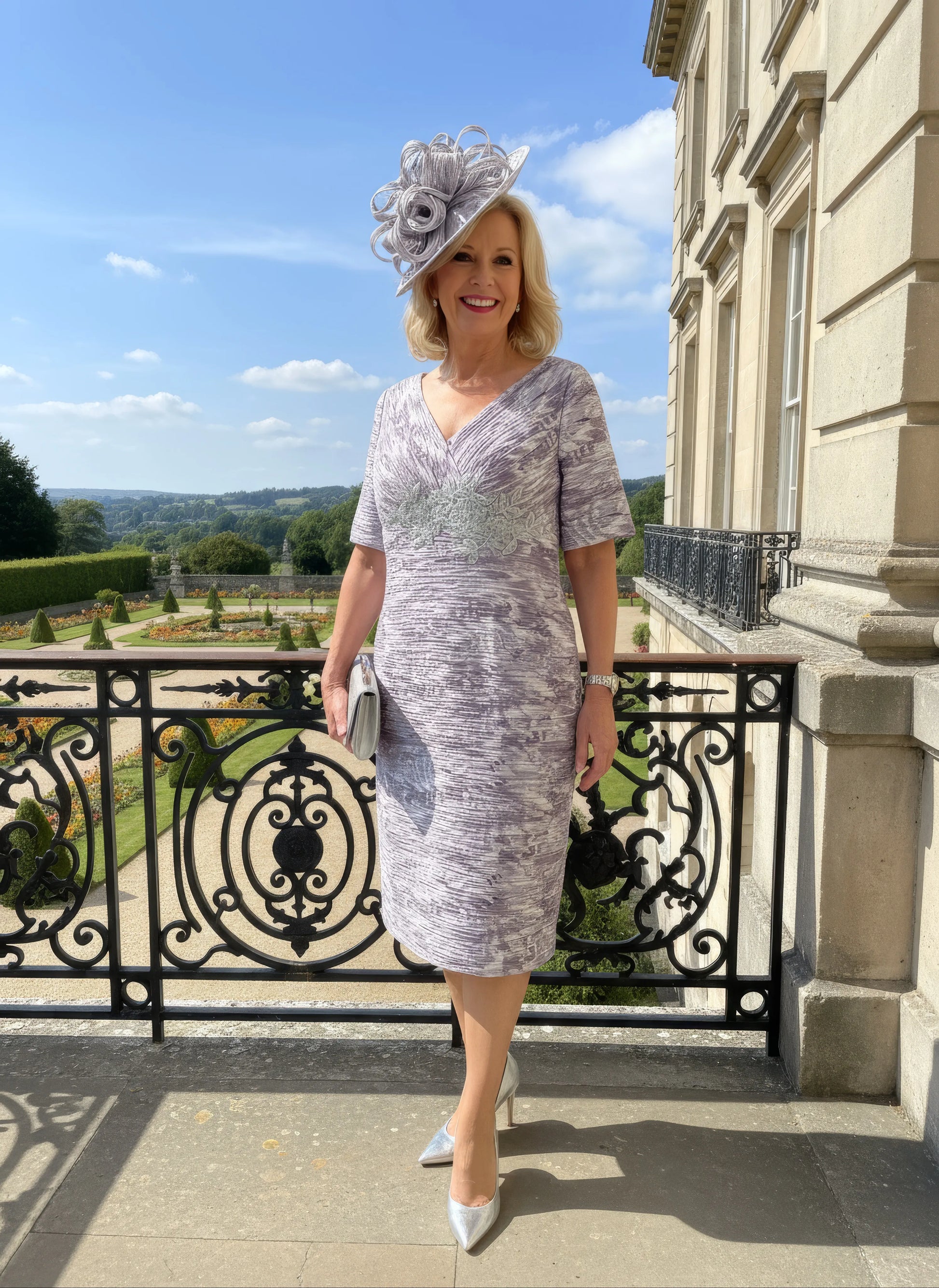 Woman in a patterned Condici grey dress as mother of the bride standing in front of a large building with greenery.