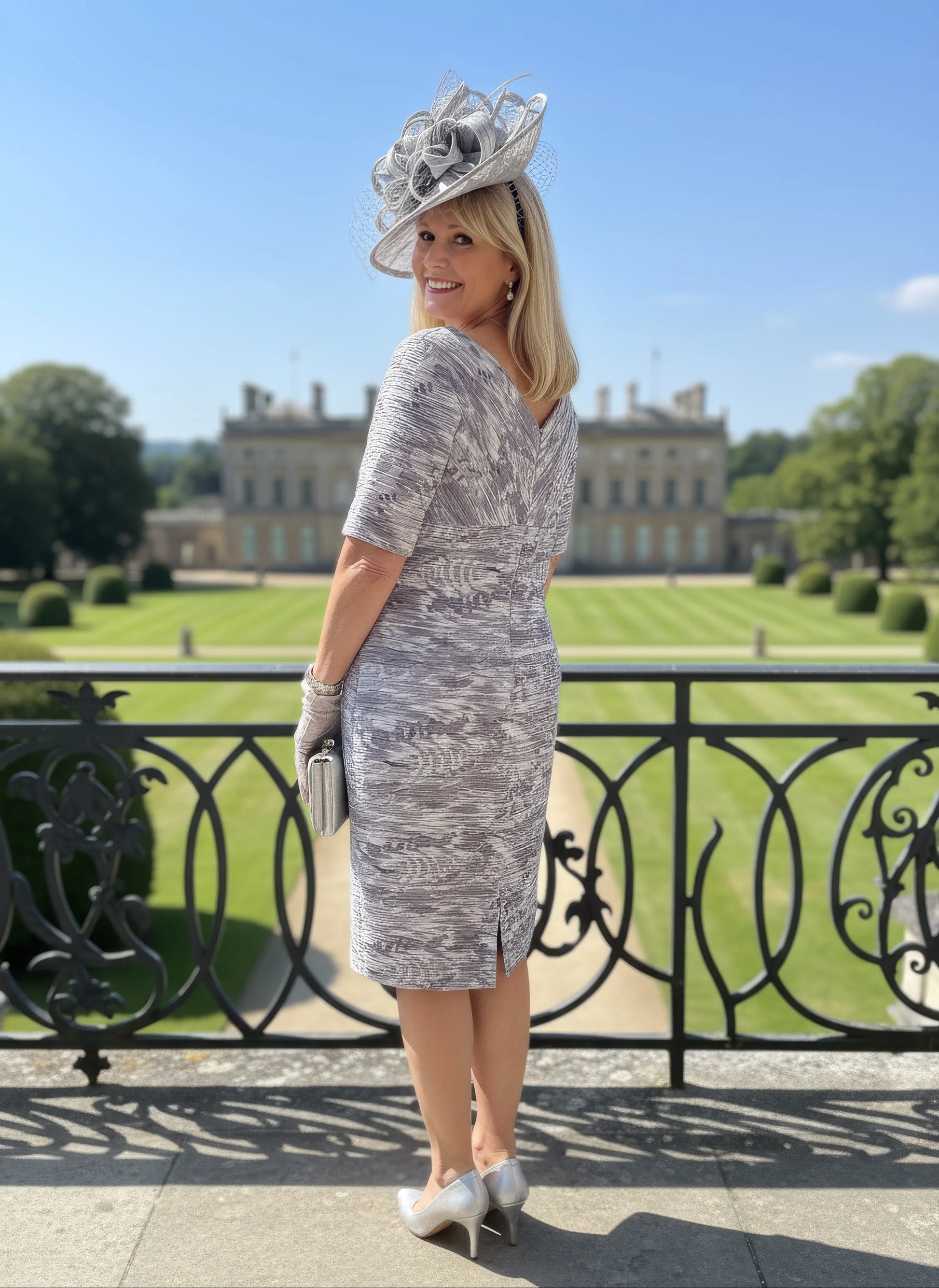 Woman in a patterned Condici grey dress as mother of the bride standing in front of a large building with greenery.