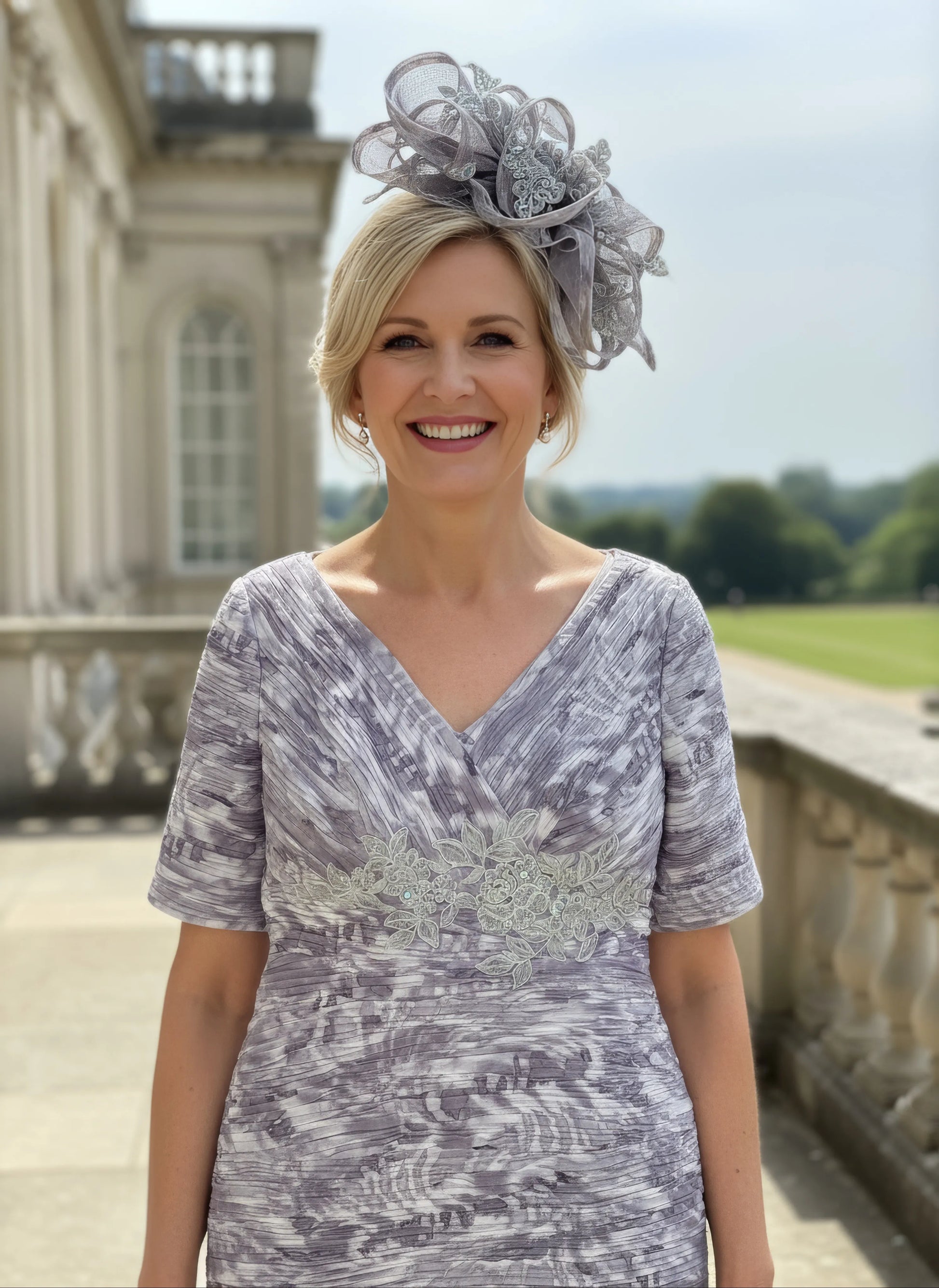 Woman in a patterned Condici grey dress as mother of the bride standing in front of a large building with greenery.