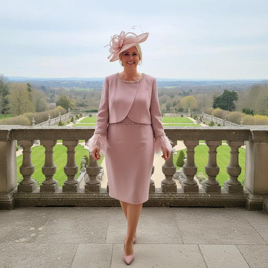 Woman in a couture club dusty pink mother of the bride outfit with a matching pink fascinator embellished hat standing on a balcony with a scenic view.