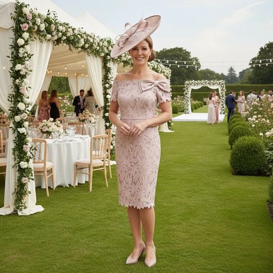Woman in a blush pink mother of the bride lace dress and matching pink fascinator standing in a garden with floral decorations and tables.