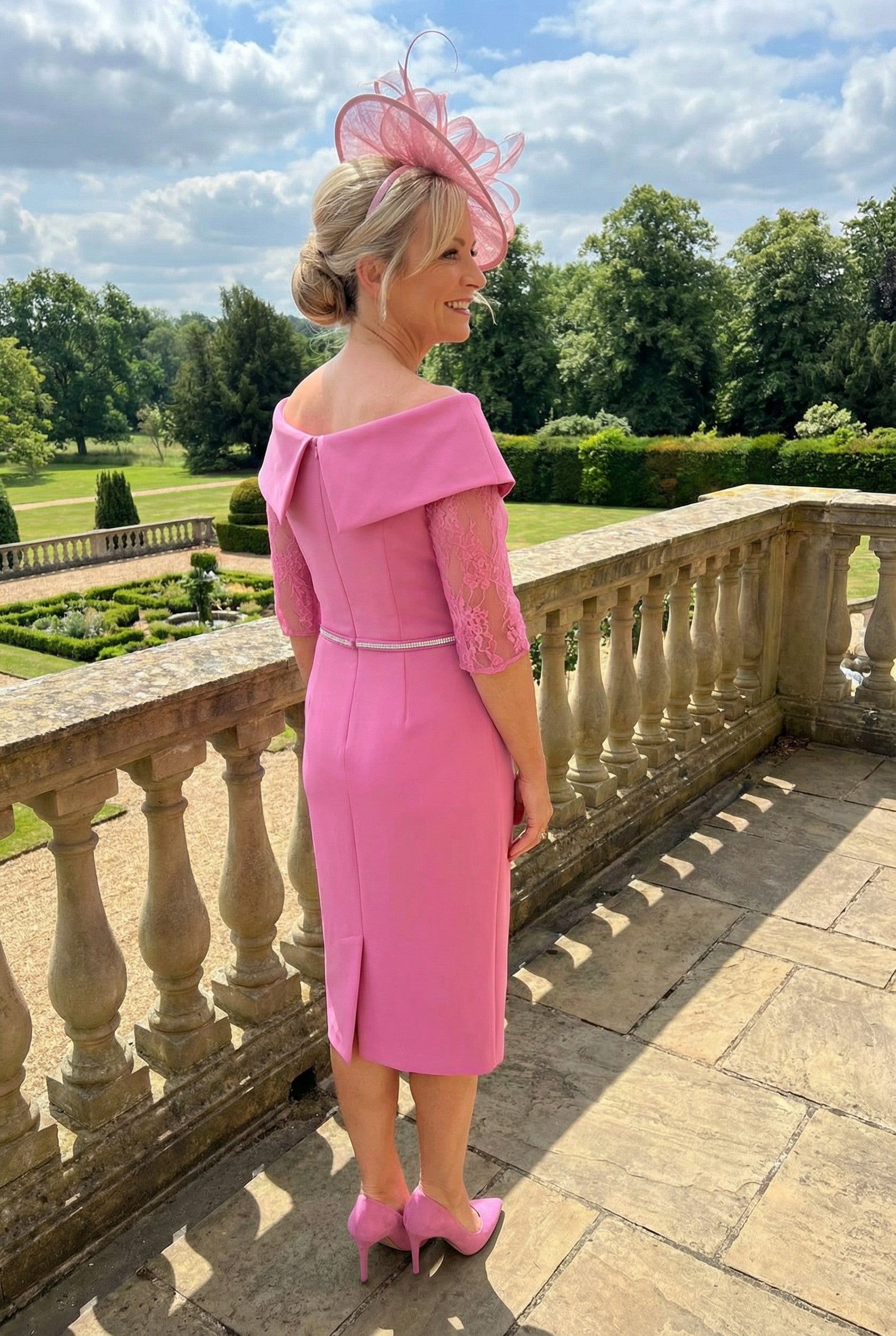 Back view of a woman in a Gabriella sanchez pink mother of the bride dress and matching pink fascinator standing on a balcony.