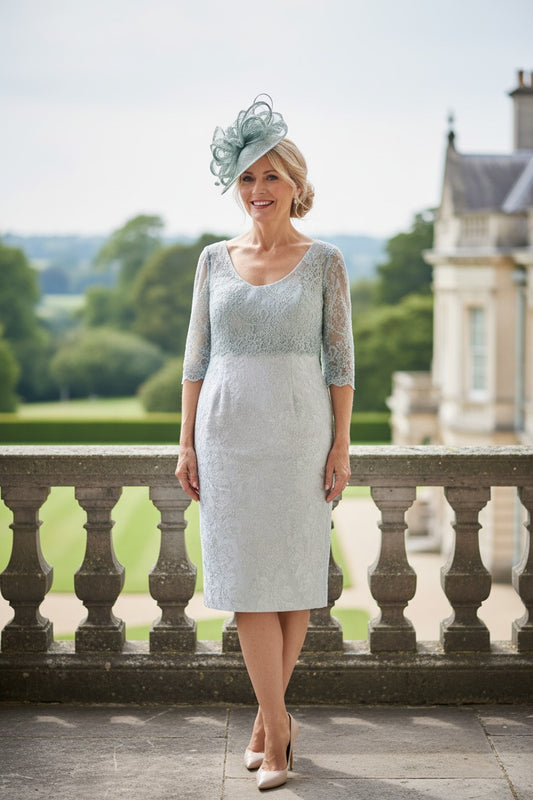 Woman in a sage green mother of the bride lace dress and sage fascinator hat standing in front of a scenic background.