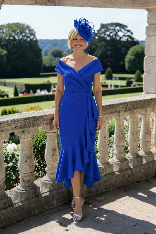Woman in a Ispirato cobalt blue mother of the bride dress andcobalt fascinator hat standing on a stone balcony with a garden view.