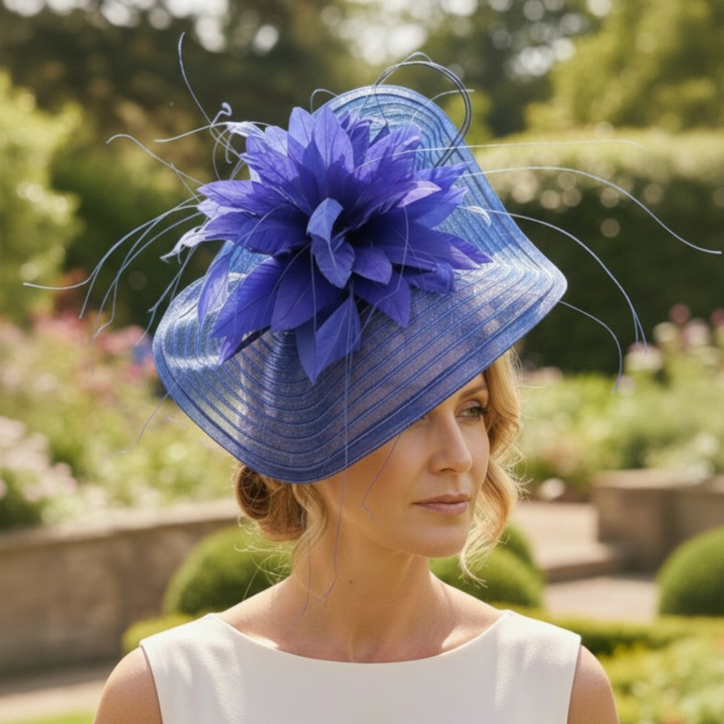Woman wearing a cobalt blue fascinator with a wide sinamay brim, large decorative flower, and curled quills, standing in a garden setting while dressed in a sleeveless white outfit