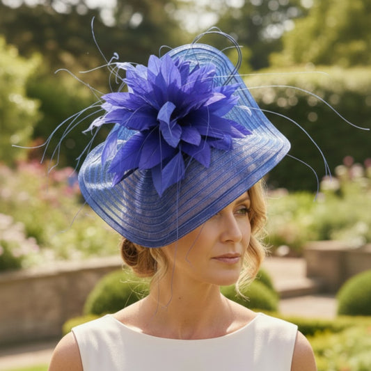 Woman wearing a cobalt blue fascinator with a wide sinamay brim, large decorative flower, and curled quills, standing in a garden setting while dressed in a sleeveless white outfit