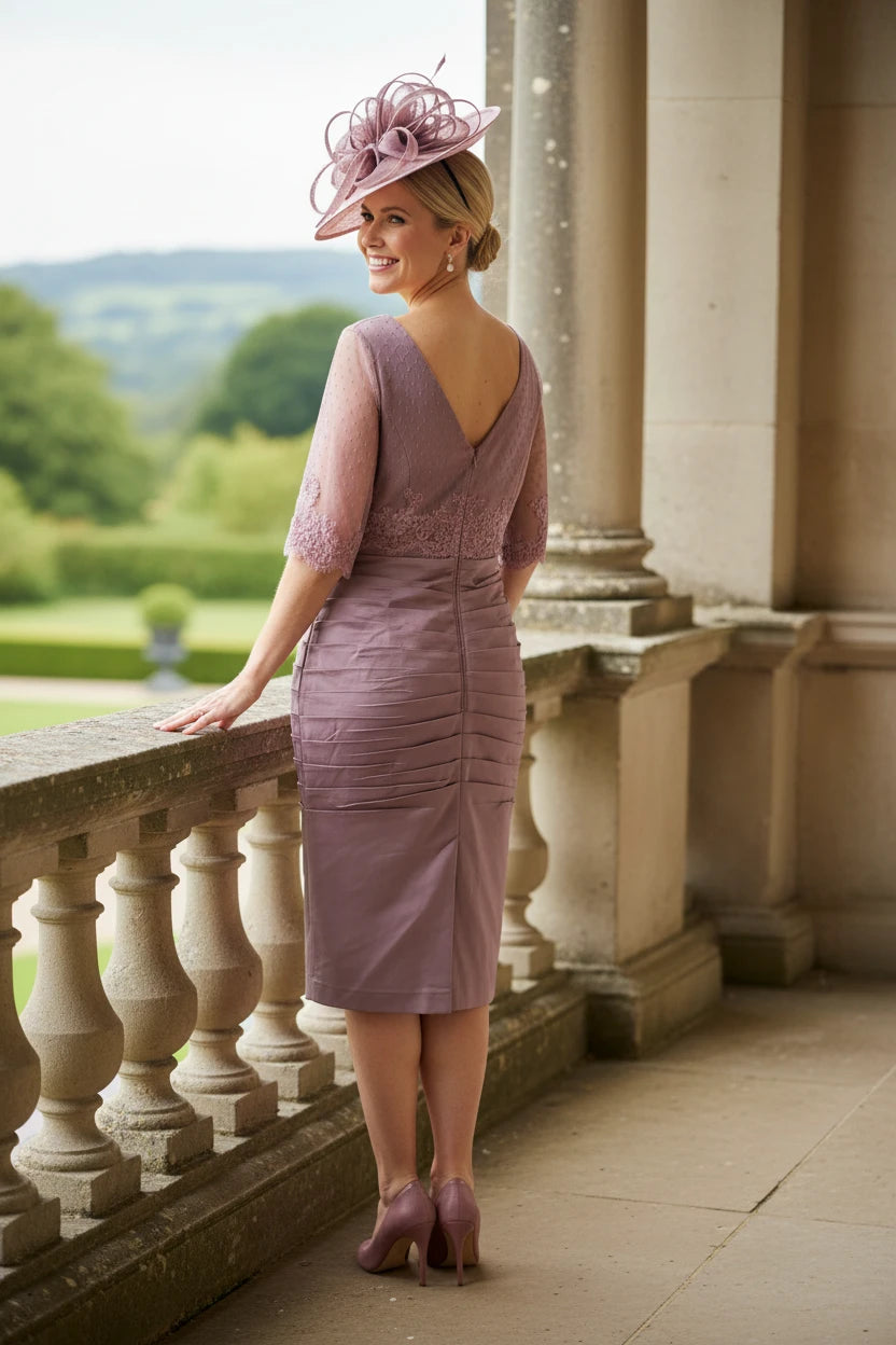 Woman in a light pink mother of the bride dress and pink fascinator hat standing on a stone balcony with a scenic background