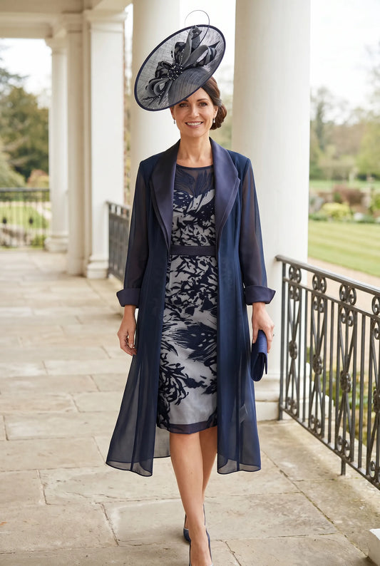 Woman in a navy mother of the bride dress with lace details and a matching coat and fascinator standing on a stone path.