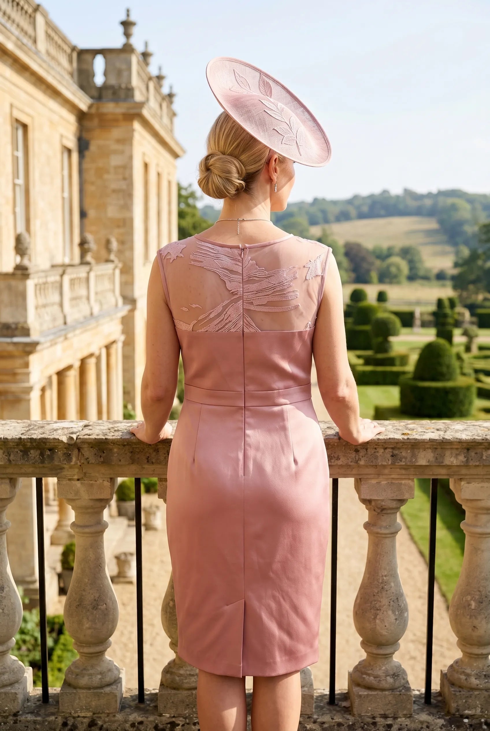 Woman in a pink mother of the bride  dress and hat standing on a balcony with a scenic background