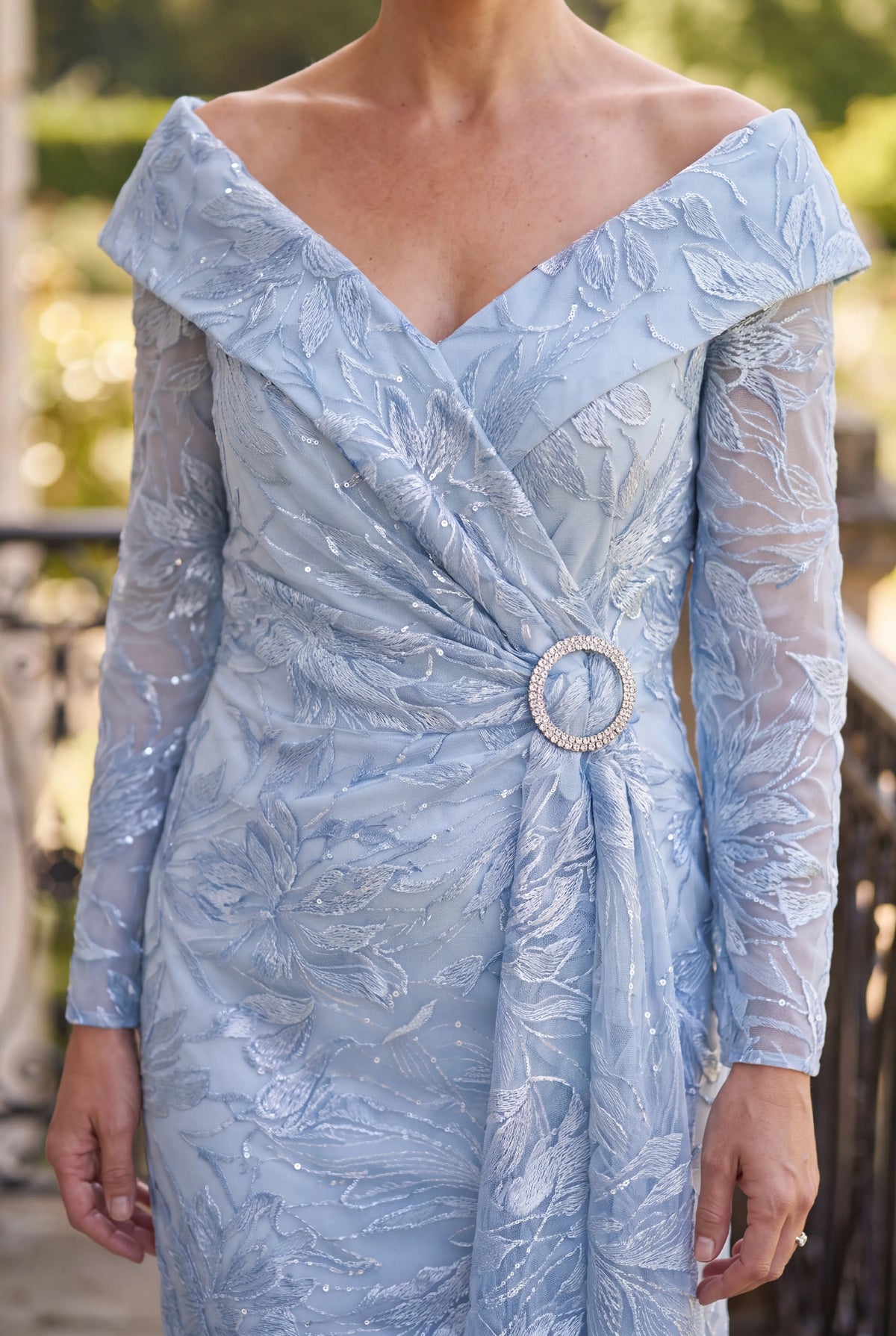 Close-up of the light blue Ispirato ISH814 Mother of the Bride dress showing its off-the-shoulder neckline, lace sleeves and decorative waist buckle.
