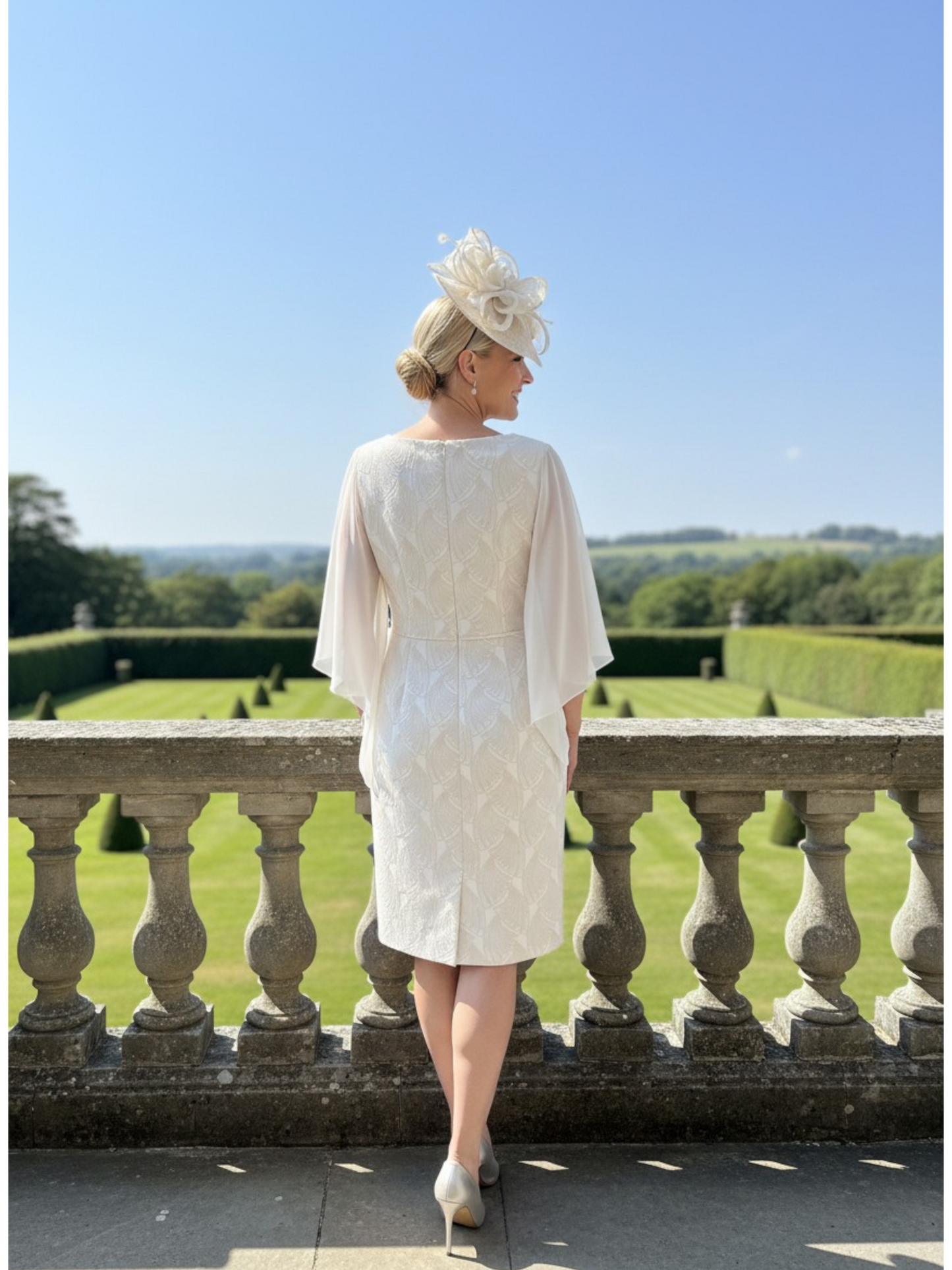 Woman in a lightweight floaty champagne mother of the bride  dress and beige fascinator hat standing on a balcony with a scenic view.