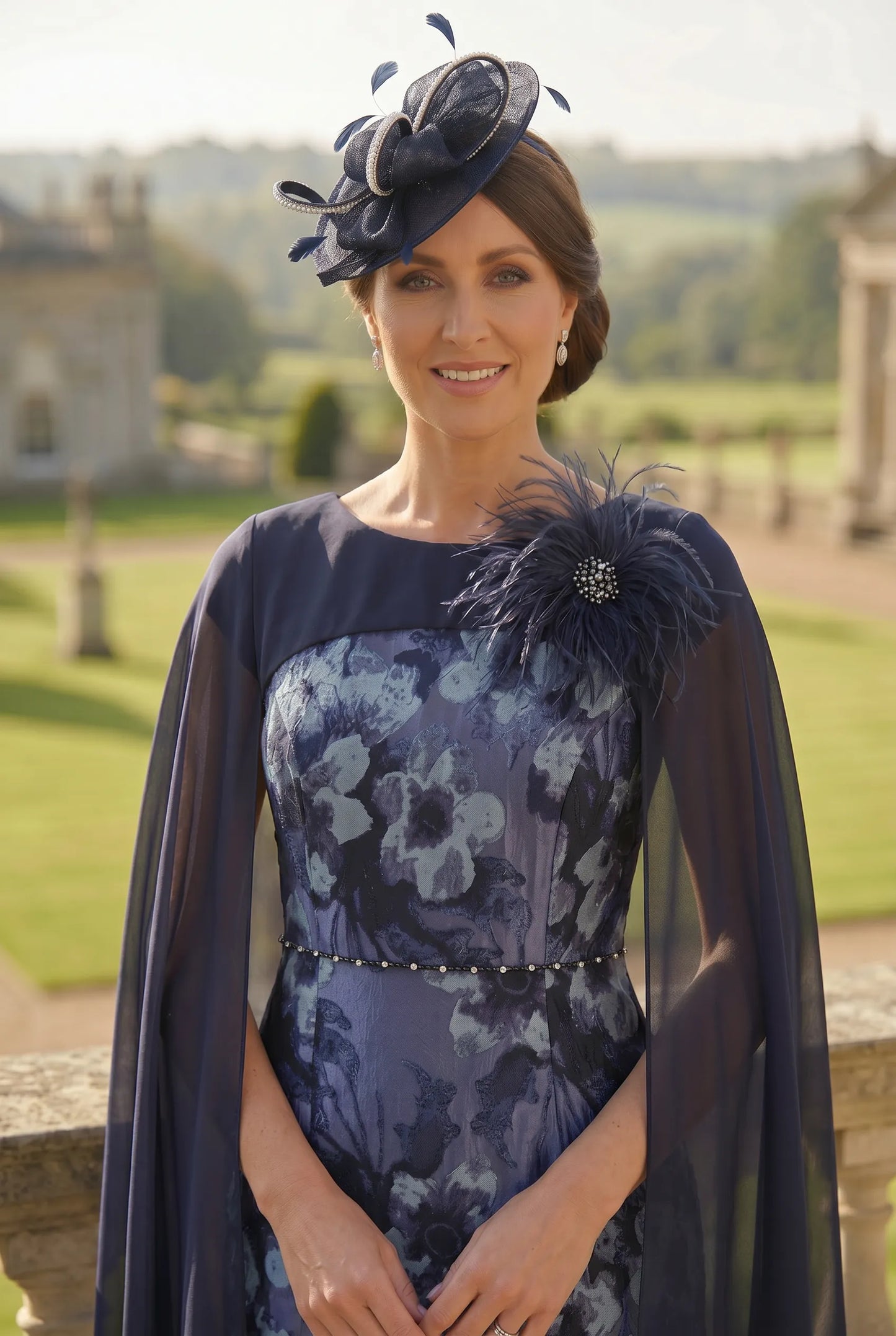 Woman wearing a dark blue floral mother of the bride dress with a matching cape and matching navy headpiece fascinator outdoors.