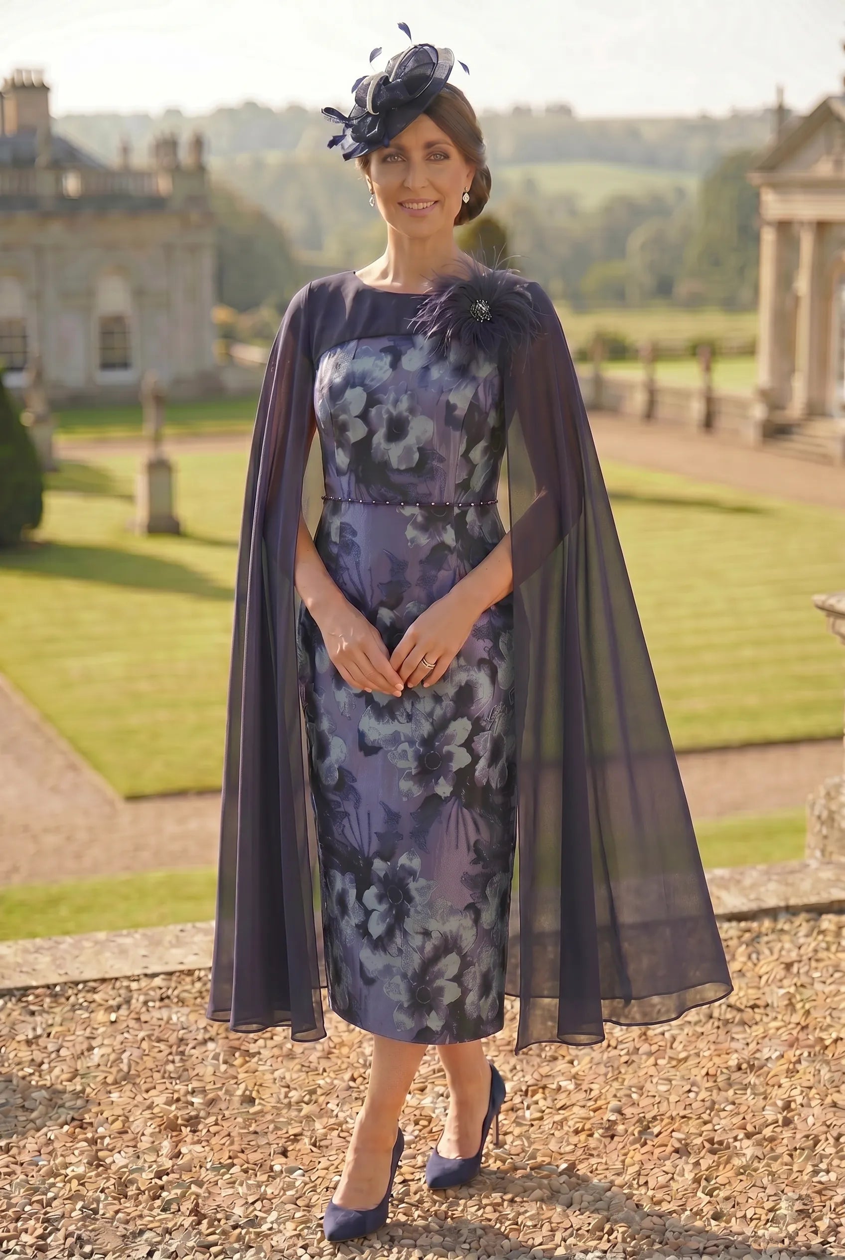 Woman in a lizabella floral mother of the bride dress with a dark navy cape standing outdoors in a garden setting.