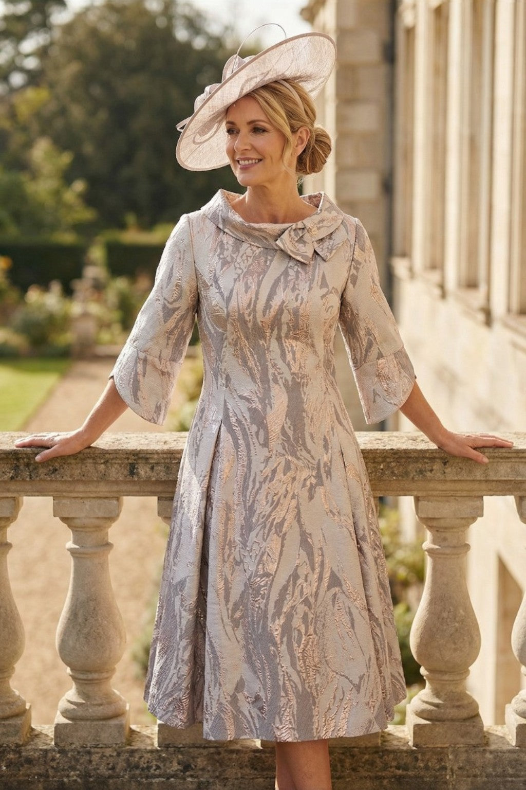 Woman in a patterned lizabella gold mother of the bride dress and champagne coloured fascinator hat standing on a stone balcony with greenery in the background