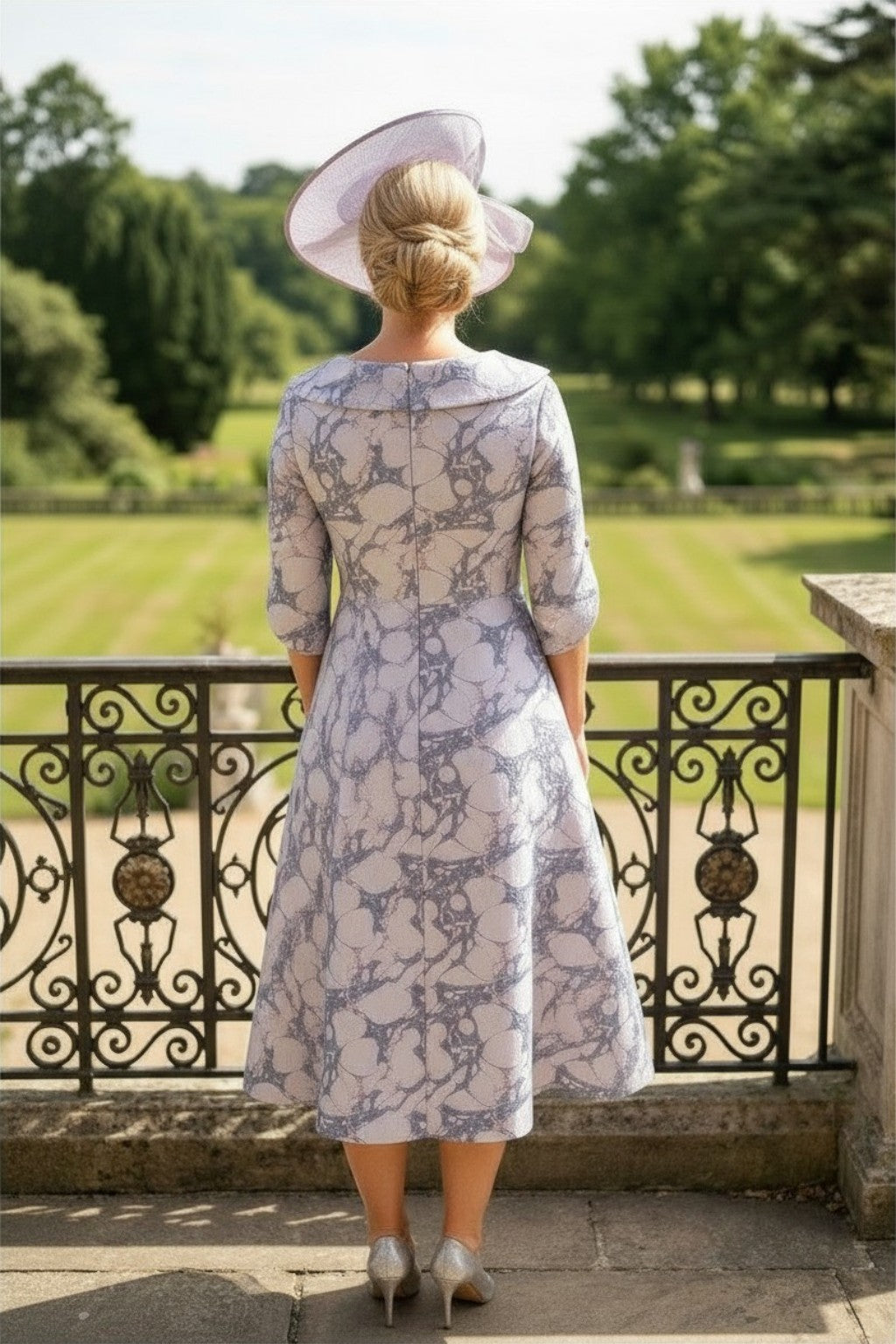 Back image of a woman is in a lizabella a-line lilac mother of the bride dress, stood on a mansion balcony with gardens in the background.