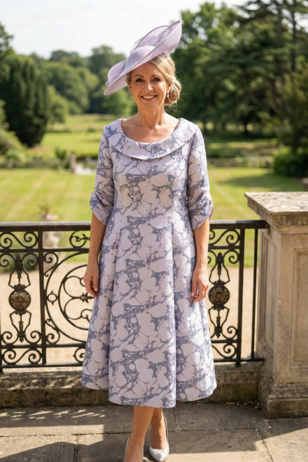 A woman is standing in a lizabella a line lilac mother of the bride dress, stood on a mansion balcony with gardens in the background.