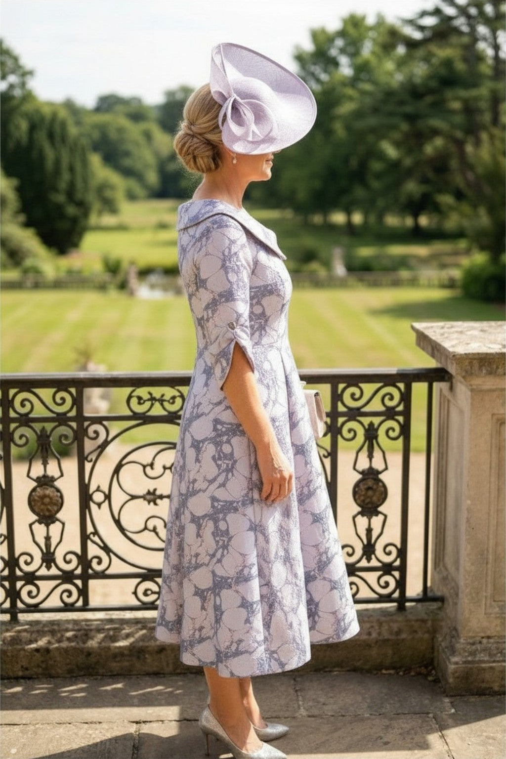 Side image of a woman is in a lizabella a-line lilac mother of the bride dress, stood on a mansion balcony with gardens in the background.