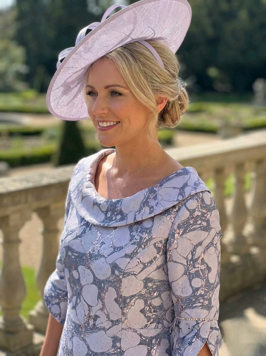 Close up image of a woman is in a lizabella a-line lilac mother of the bride dress, stood on a mansion balcony with gardens in the background.