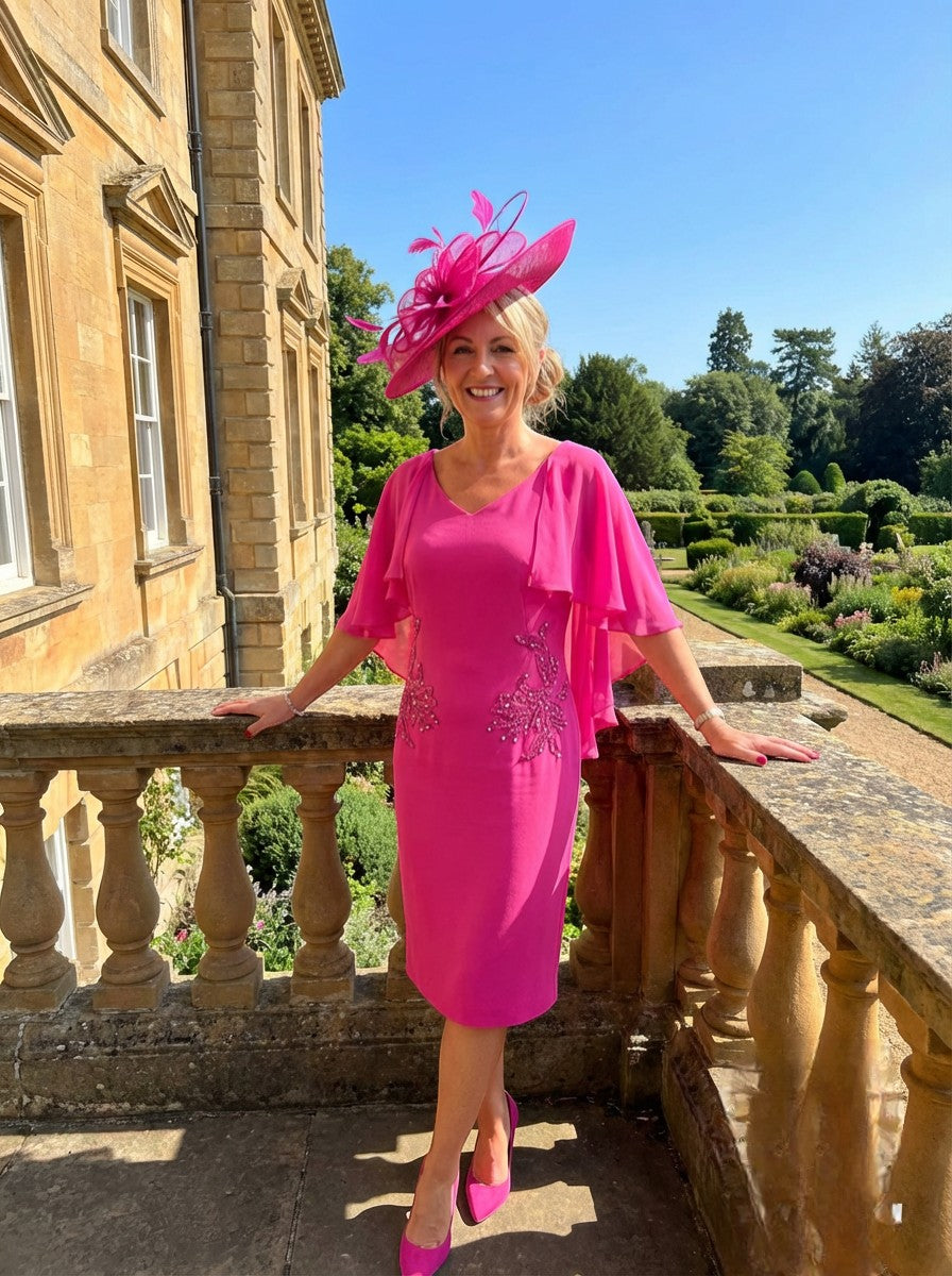 Woman in a Lizabella bright pink mother of the bride dress with chiffon cape sleeves and matching fascinator  hat standing on a stone balcony with a garden view.