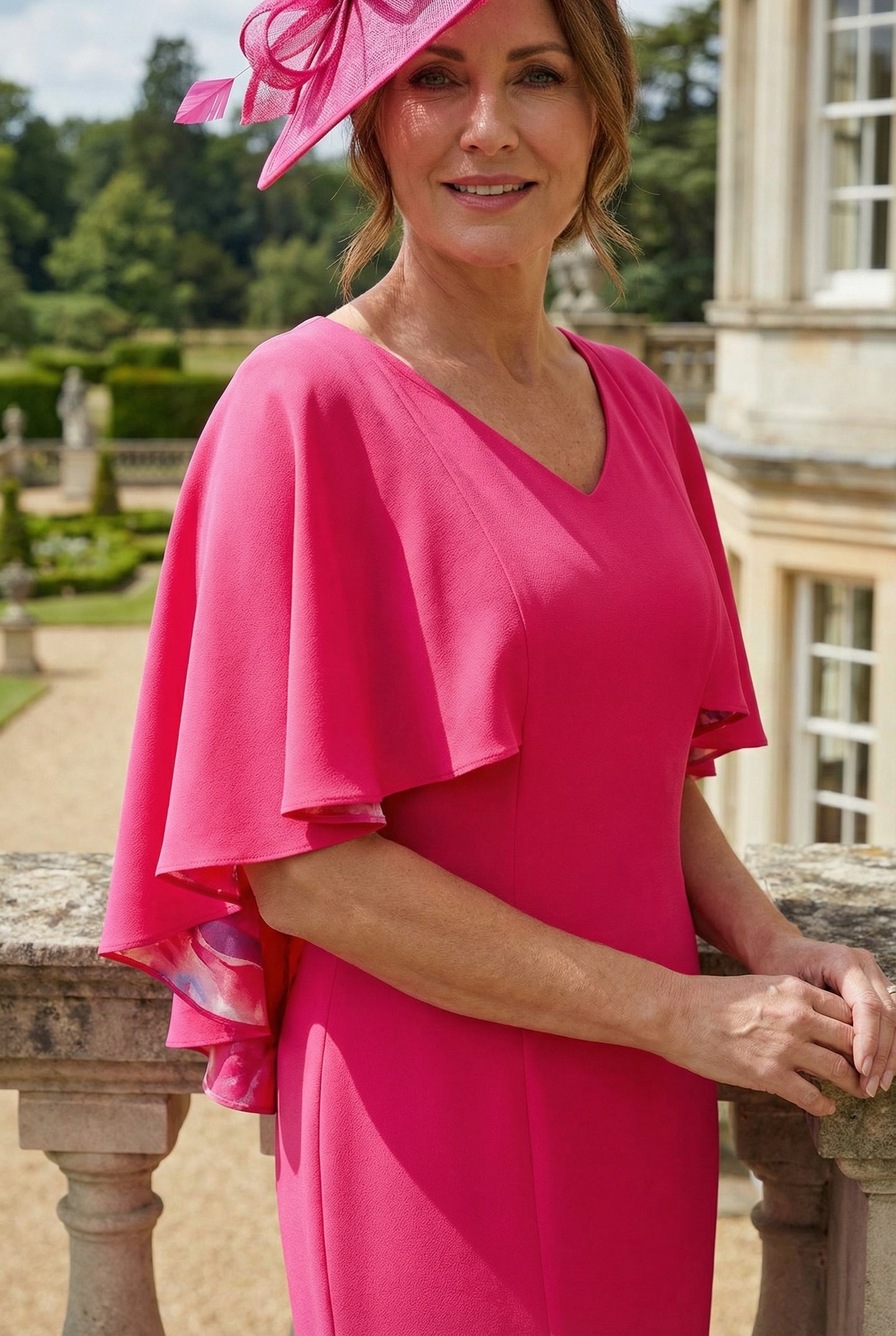 Close view of a woman in a Lizabella bright pink mother of the bride dress with ruffled sleeves and matching fascinator  hat standing on a stone balcony with a garden view.