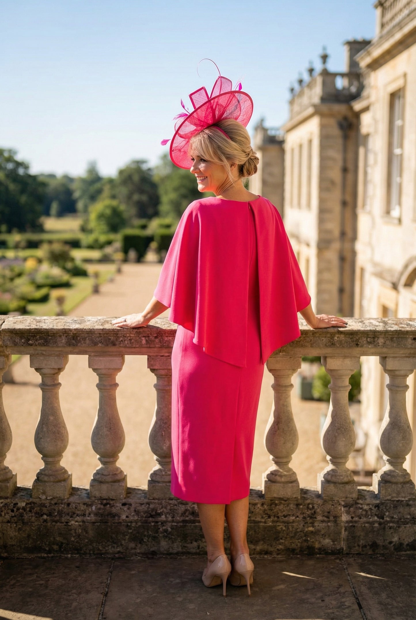 Back view of a woman in a Lizabella bright pink mother of the bride dress with ruffled sleeves and matching fascinator  hat standing on a stone balcony with a garden view.