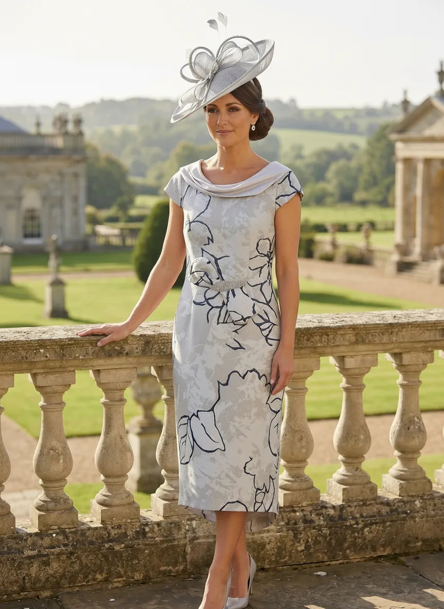 Woman in a silver mother of the bride  dress and silver fascinator hat standing on a stone balcony with a scenic. background