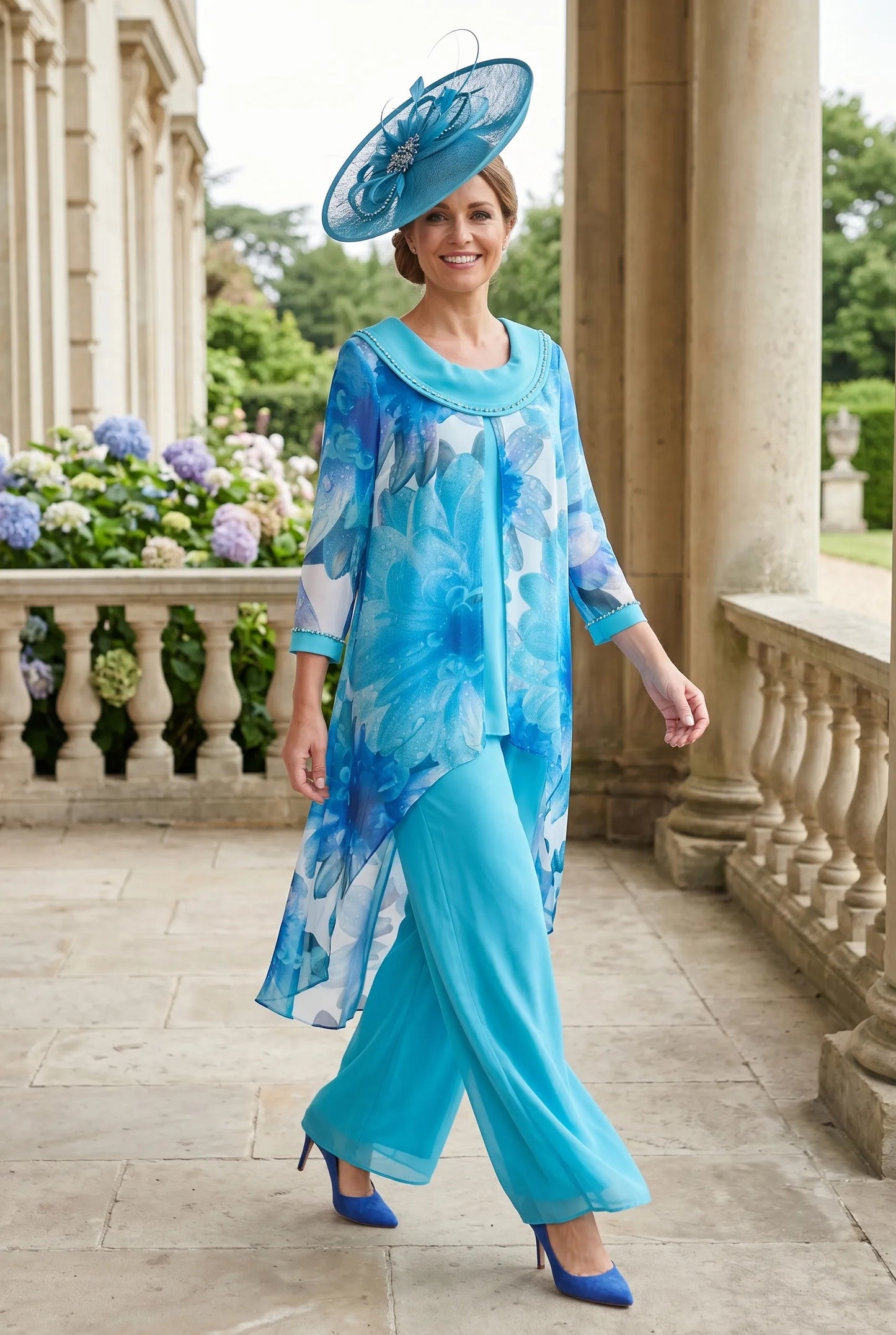 Woman in a blue floral mother of the bride 2 piece outfit and wearing a blue embellished fascinator wedding hat standing on a stone path with decorative columns and flowers in the background.