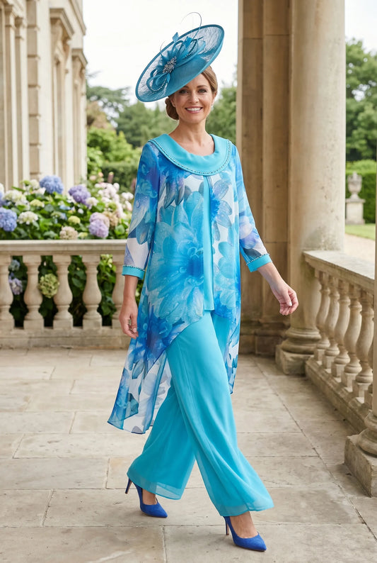 Woman in a blue floral mother of the bride 2 piece outfit and wearing a blue embellished fascinator wedding hat standing on a stone path with decorative columns and flowers in the background.