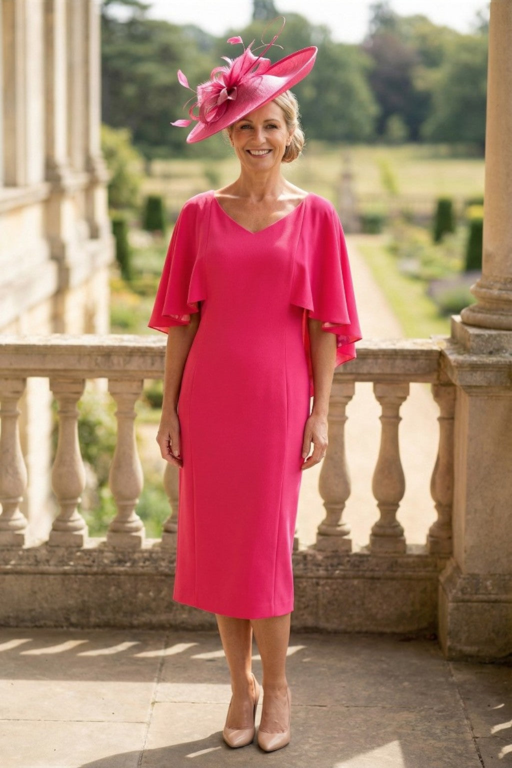 Woman in a Lizabella bright pink mother of the bride dress with ruffled sleeves and matching fascinator  hat standing on a stone balcony with a garden view.