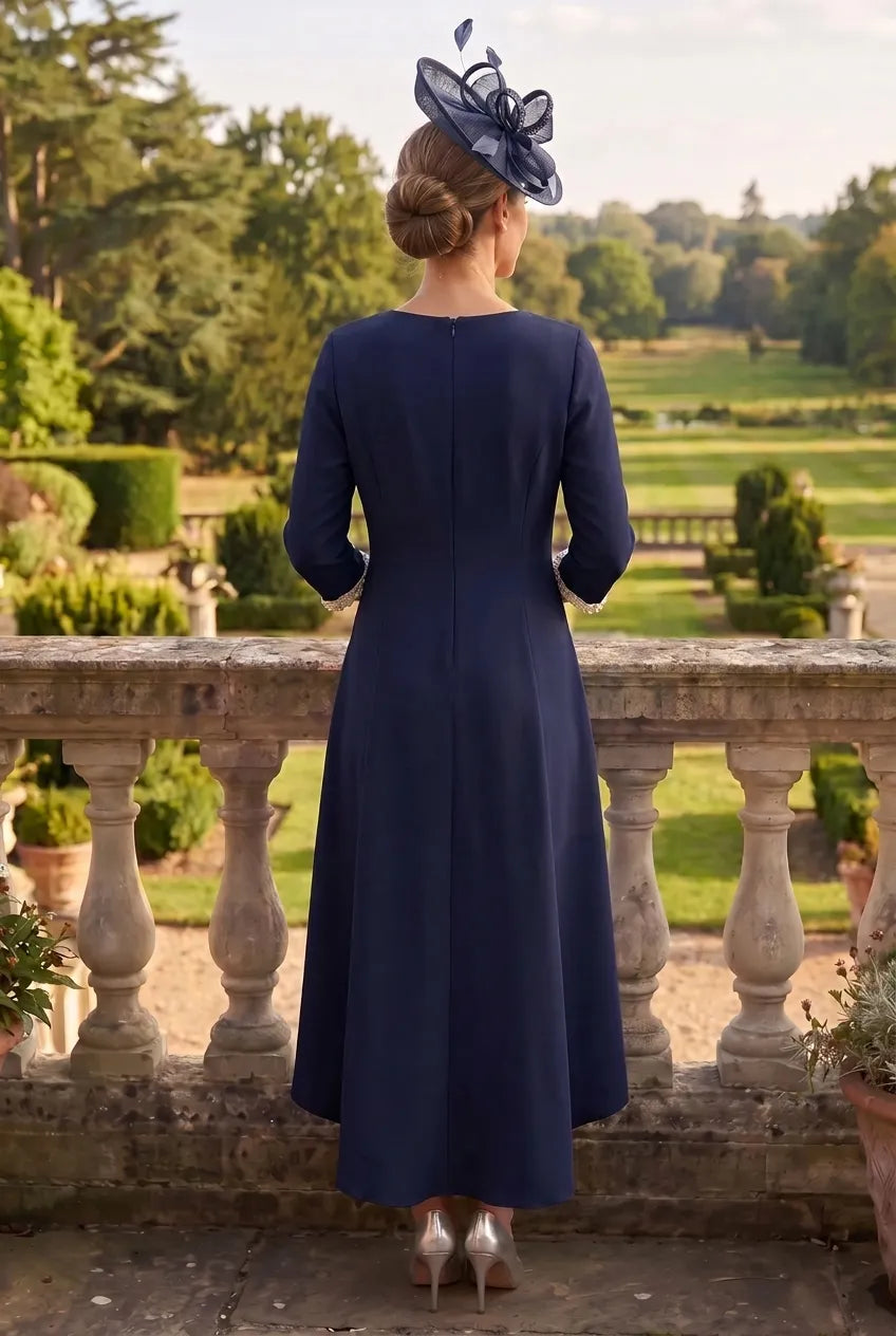 A woman stands with her back to the camera wearing a navy blue long-sleeve midi dress and a matching fascinator hat in an outdoor garden setting.
