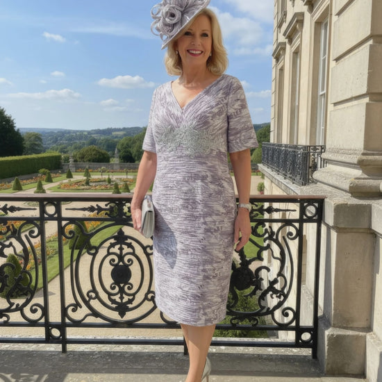 Woman in a patterned Condici grey dress as mother of the bride standing in front of a large building with greenery.