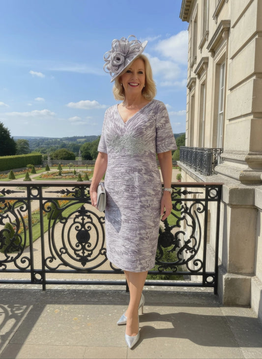 Woman in a patterned Condici grey dress as mother of the bride standing in front of a large building with greenery.