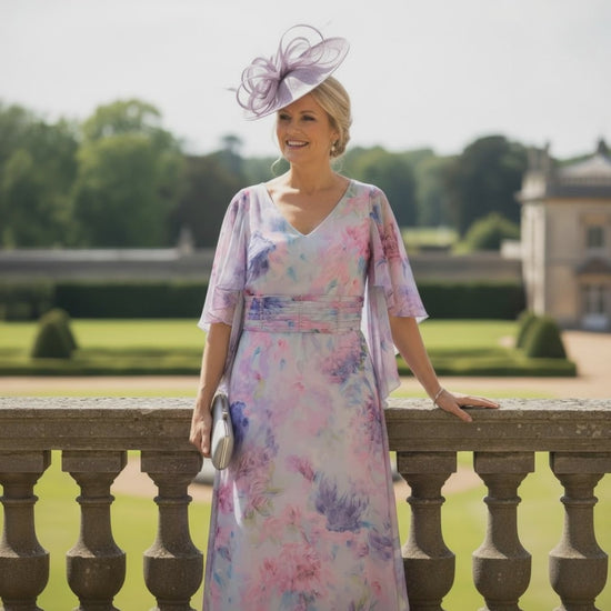 Woman in lizabella floral lavender mother of the bride dress and hat standing in a garden setting. Signature Dress