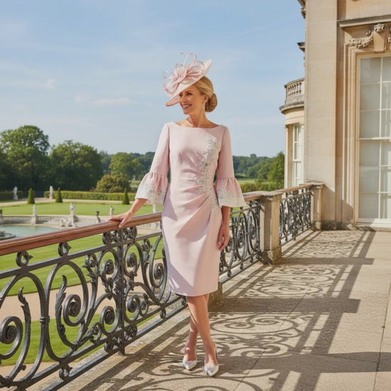 Woman in a light blush pink John Charles mother of the bride dress and matching pink fascinator hat standing on a balcony with a scenic background. Signature Dress.