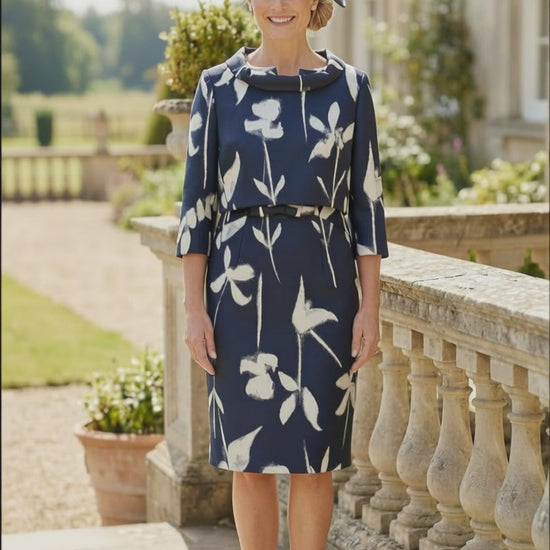 A video of a Navy floral mother of the bride dress with matching navy fascinator, walking on a stone terrace overlooking a formal garden.
