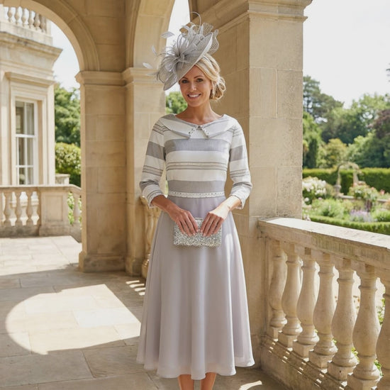 Woman in a Lizabella striped A Line mother of the bride dress with a matching silver fascinator hat standing on a balcony with classical architecture. Signature Dress.
