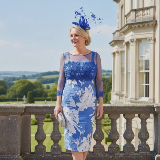 Woman in a cobalt blue mother of the bride dress with a matching jacket and headpiece, standing in front of a classical building. Signature Dress