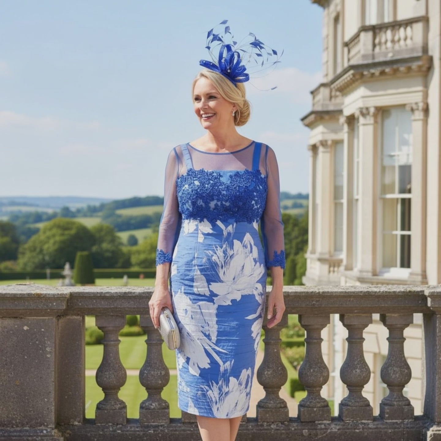 Woman in a cobalt blue mother of the bride dress with a matching jacket and headpiece, standing in front of a classical building. Signature Dress