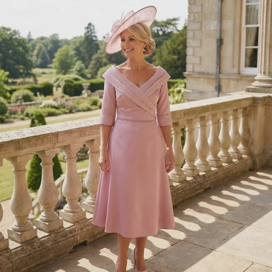 Lizabella pink midi dress with pleated neckline and matching pink fascinator, worn on a balcony overlooking formal gardens.